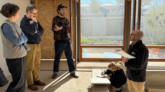 Four adults discussing plans inside a home under construction, beside large timber-framed windows, with a child drawing nearby.