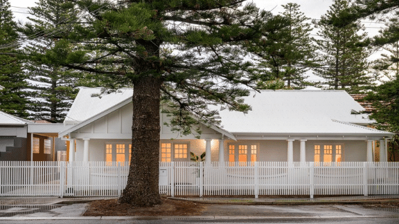 White character home with a pitched metal roof, warm-lit windows and a white picket fence, framed by tall pine trees.